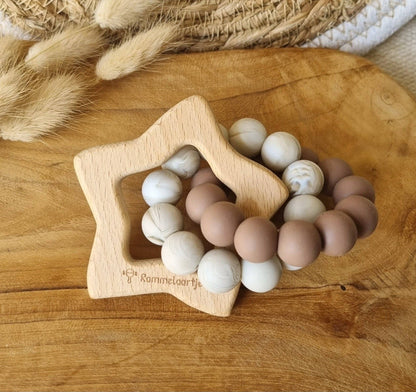 Beißring mit Holzstern und braun-beigen Silikonperlen auf Holzbrett, daneben Trockengras
