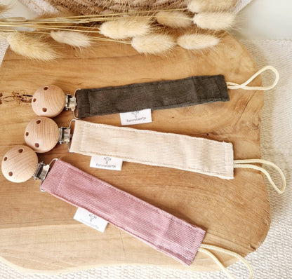 Drei Cord-Schnullerhalter in Army-Grün, Beige und Puderrosa mit Holzclips nebeneinander auf Holzbrett