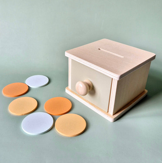 Helle Holz-Spielbox mit Schublade und Holzknauf, davor sechs Scheiben in Orange, Gelb und Weiß auf grünem Hintergrund.