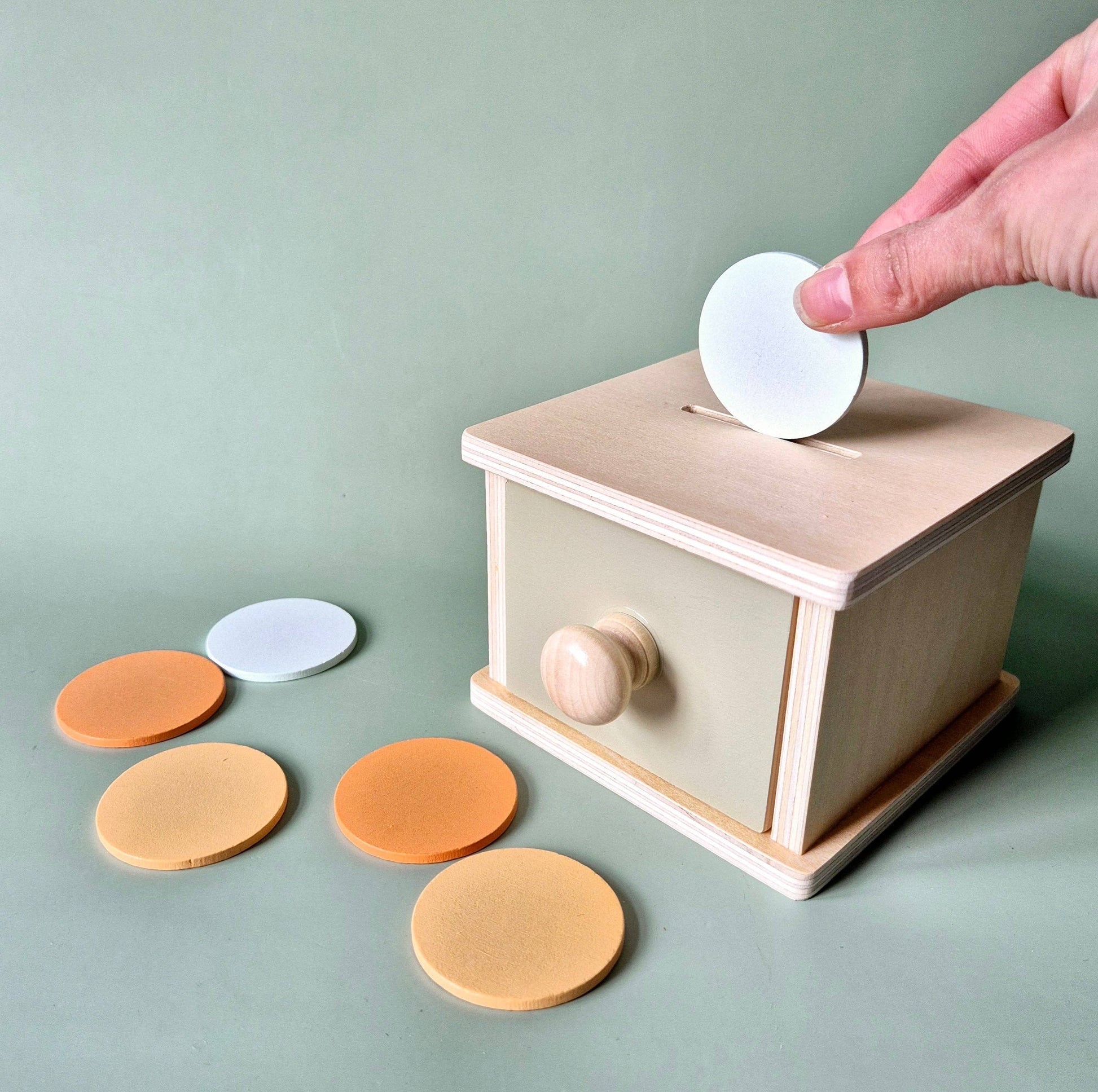 Nahaufnahme: Hand führt eine weiße Scheibe in den Schlitz der Holzbox, orange und gelbe Scheiben liegen daneben.