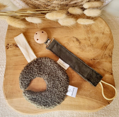 Schnullerhalter in Army-Grün neben grauem Greifring aus Bouclé und Holzclip auf Holzbrett
